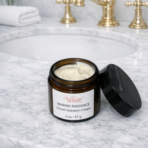 Open jar of Velvet Marine Radiance Cloud Hydration Cream resting on a bright white marble bathroom countertop beside a gold faucet and folded towel. The amber jar features a white label with rose‑gold Velvet logo and black text, showing the cream’s smooth, whipped texture inside.