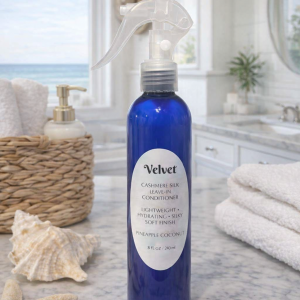 Velvet Cashmere Silk Leave‑In Conditioner in a cobalt blue spray bottle displayed on a marble countertop in a bright beach‑house bathroom. The bottle sits beside seashells, a starfish, and rolled white towels with sunlight streaming through a window overlooking the ocean