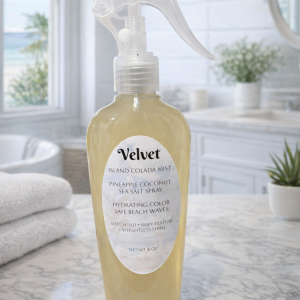 Velvet Island Colada Mist sea salt spray bottle on a white marble counter in a bright beach‑house bathroom, with soft natural light and coastal décor in the background.