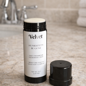 A black Velvet “Humidititty Be Gone” underboob deodorant stick with its cap removed, displayed on a beige bathroom countertop beside a chrome faucet and folded white towels.